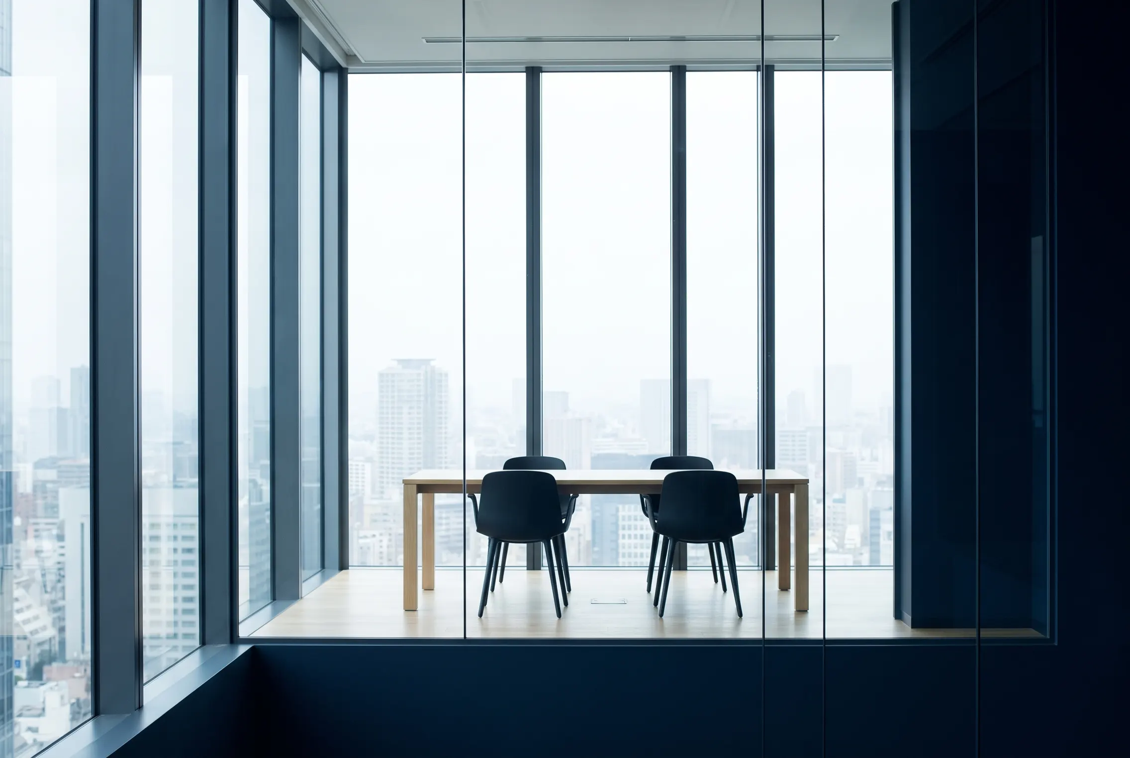 A glass-walled meeting room high above an overcast city, with a slim table and two chairs